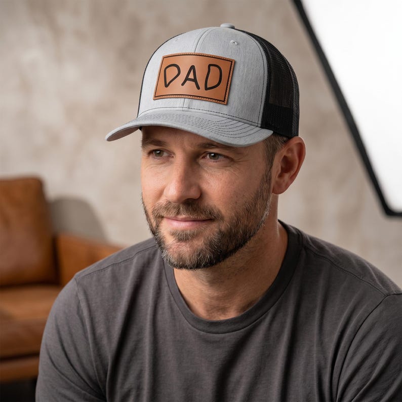 May include: A gray and black trucker hat with a leather patch that reads "DAD" in black lettering. The hat has a curved brim and a mesh back. The hat is a classic style, suitable for casual wear.