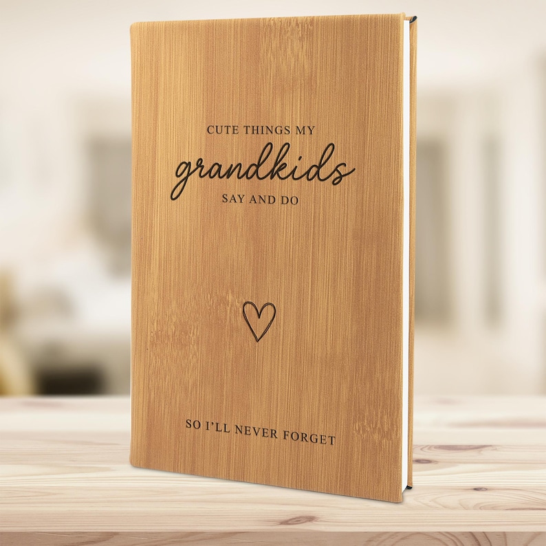 K&ouml;nnte beinhalten: Ein Notizbuch mit hellbraunem Bambuseinband, auf dessen Vorderseite der eingravierte Text "CUTE THINGS MY grandkids SAY AND DO", ein kleines Herzsymbol und der Satz "SO I'LL NEVER FORGET" mit sichtbarer Holzmaserung zentriert sind.