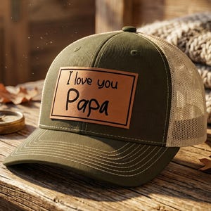 May include: Olive green and tan trucker hat with the text "I love you Papa" on a brown leather patch. The hat has a curved brim and a mesh back, perfect for outdoor wear.