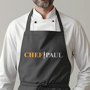 May include: A black apron featuring the text CHEF PAUL in orange and white lettering, with a white chef knife icon replacing the letter A. The apron is worn over a white long-sleeved chef coat with a mandarin collar and black buttons.