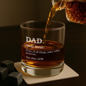 Puede incluir: Un vaso transparente con la palabra "DAD" grabada en la parte delantera, lleno de l&iacute;quido &aacute;mbar. El texto de abajo define "DAD" como un sustantivo, enumerando cualidades como "reparador" y "protector". Se est&aacute; vertiendo whisky en el vaso.