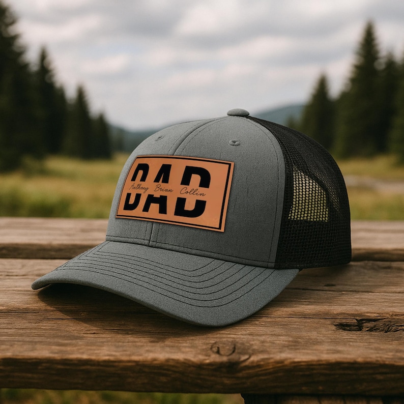 K&ouml;nnte beinhalten: Graue Trucker-Cap mit schwarzem Mesh-R&uuml;cken und gebogenem Schirm. Die Kappe hat einen braunen Lederaufn&auml;her mit dem Wort "DAD" in fetten schwarzen Buchstaben und den Namen "Anthony Brian Collin" in kleinerer Schrift.
