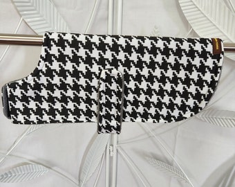 May include: Black and white houndstooth dog coat with a gray strap. The coat is hanging on a white metal stand.