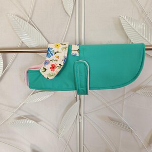 May include: A teal dog coat featuring a floral patterned collar and pink lining. The coat has a curved back and a strap with white trim. The floral pattern includes blue, yellow, and red flowers on a white background.