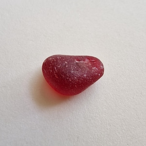 May include: A piece of red sea glass, shaped like a heart. The glass has a frosted texture and an uneven form. The colour is a deep, rich red, with some tonal variations. This sea glass likely originates from a bottle or other glass item.