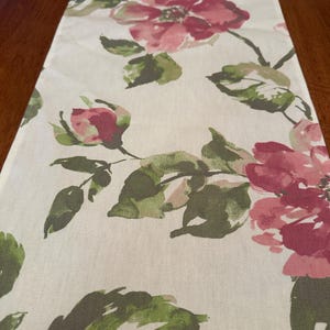 May include: A white table runner with a pink and green floral pattern. The flowers are painted in a watercolor style and have a soft, romantic look.