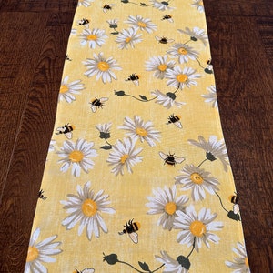 May include: A yellow table runner with a white daisy and bee pattern. The runner is made of a soft, textured fabric.
