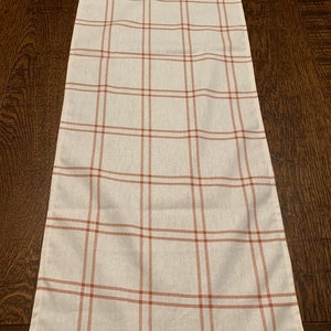 May include: A white table runner with a red and white plaid pattern. The runner is made of a soft, woven fabric and has a subtle sheen.