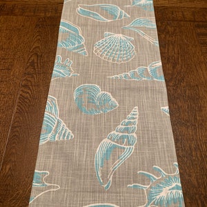 May include: A gray and white table runner with a blue seashell pattern. The seashells are in various shapes and sizes, and some have a brown outline.