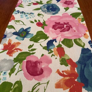 May include: A white table runner with a floral pattern. The flowers are painted in shades of pink, blue, orange, and green. The runner is perfect for adding a touch of color and elegance to any table.