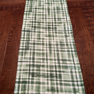 May include: A green and white plaid table runner with a watercolor effect. The runner is made of fabric and has a subtle sheen.
