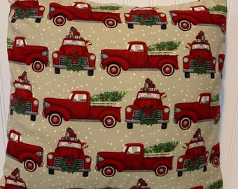 May include: A beige throw pillow with a red truck pattern. The trucks are carrying Christmas trees and presents. The background is a light beige with white dots.