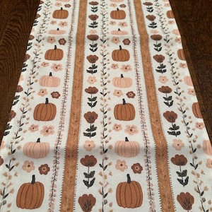 May include: A fall-themed table runner featuring a repeating pattern of pumpkins and floral elements in shades of brown, peach, and cream. The design includes alternating rows of pumpkins and flowers, with vertical stripes of a textured pattern. This runner is perfect for seasonal home decor.