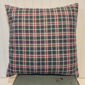 May include: A gray and red plaid throw pillow with a square pattern. The pillow is sitting on a gray metal surface.