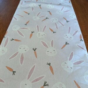 May include: A white table runner with a pink and white bunny and carrot pattern. The runner is made of a soft, woven fabric.