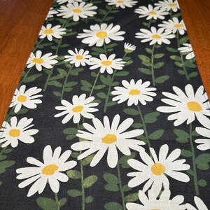 May include: A black table runner with a white daisy pattern. The daisies have yellow centers and green stems.
