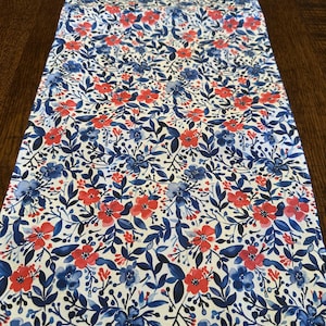 May include: A white table runner with a red and blue floral pattern. The pattern features small red flowers and blue leaves on a white background.