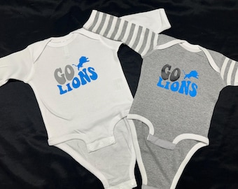 May include: Two baby onesies, one white and one gray, with the words "Go Lions" in blue and a blue lion graphic. The gray onesie has white stripes on the sleeves.