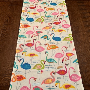 May include: A white table runner with a repeating pattern of colorful flamingos wearing sunglasses. The flamingos are standing in a wavy pattern that resembles water.
