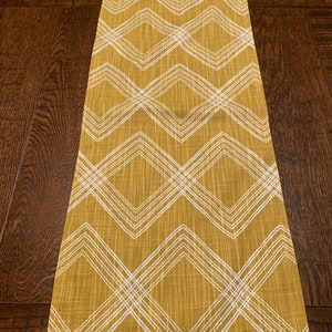 May include: Mustard yellow table runner with a white geometric diamond pattern. The runner is rectangular and made of fabric. The design features intersecting lines creating a series of diamond shapes. The runner is placed on a dark brown wooden surface.