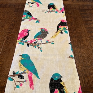 May include: A white table runner with a colorful bird pattern. The birds are blue, green, yellow, pink, and black. The runner is made of a linen fabric.