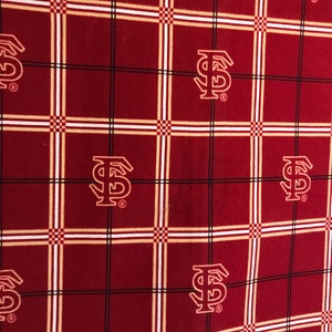May include: Maroon fabric with a plaid pattern featuring the letter "F" and "S" in a stylized design. The plaid includes white, black, and gold lines, creating a grid-like appearance. The repeating logo adds a collegiate or team-themed aesthetic.