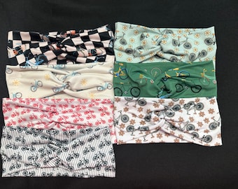 May include: Six different patterned headbands with a twist design. The headbands feature a variety of bicycle designs, including black and white checkered, floral, and striped patterns.