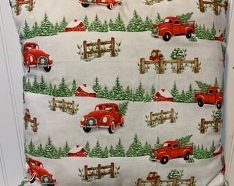 May include: A white pillow with a red truck and Christmas tree pattern. The trucks are carrying Christmas trees and are driving through a snowy winter scene. There are also small red houses and fences in the pattern.