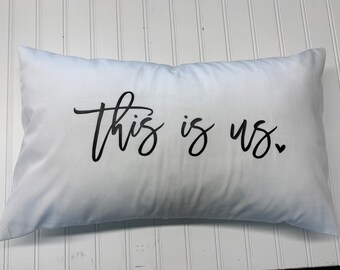 May include: White pillow with black cursive text that reads "This is us." with a small heart after the "us".