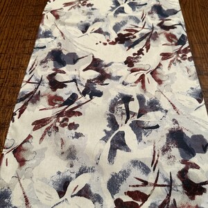 May include: A white table runner with a floral pattern in shades of burgundy, gray, and blue. The pattern features abstract leaves and flowers.