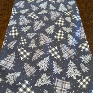 May include: A dark blue Christmas table runner featuring a repeating pattern of stylized Christmas trees. The trees are in various designs, including plaid, leopard print, and solid white, with a scattering of small white dots.