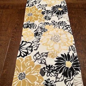 May include: A white table runner with a floral pattern in black, gold, and gray. The flowers are large and bold, and the runner is perfect for adding a touch of elegance to any table.