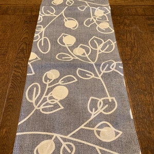 May include: A blue and white table runner with a floral pattern. The runner features a repeating pattern of white leaves and flowers on a blue background.