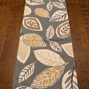 May include: A gray table runner with a leaf pattern in shades of brown, beige, and white. The leaves are stylized and have a modern design.