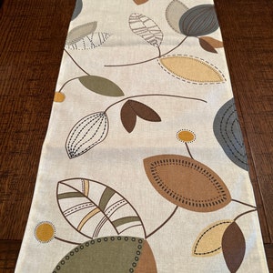 May include: A beige table runner with a leaf pattern in shades of brown, green, and yellow. The leaves are stylized and have a stitched look.