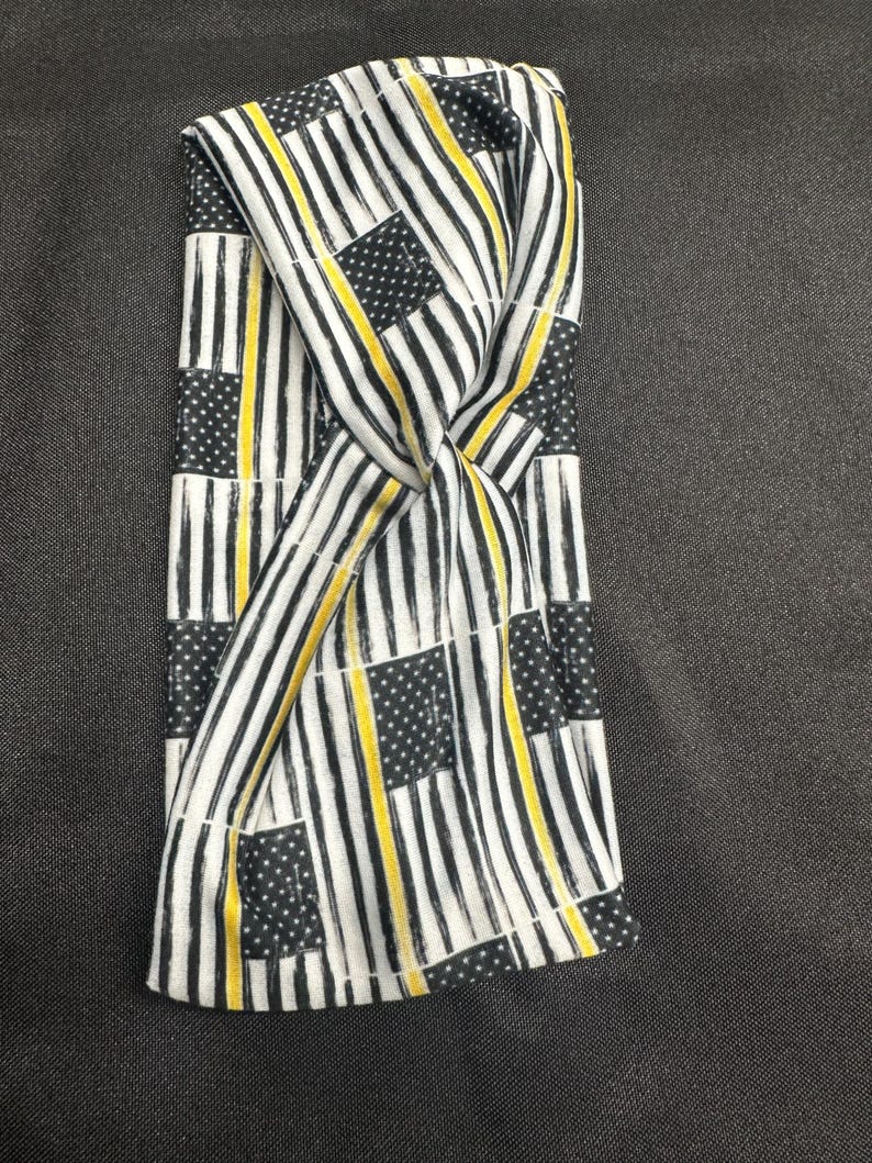 May include: A black and white headband with a thin yellow stripe pattern. The pattern is a repeated design of American flags with a yellow stripe across the middle.