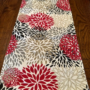 May include: A white table runner with a red, black, and gray floral pattern. The runner is made of fabric and has a subtle sheen.