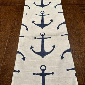 May include: A white table runner with a navy blue anchor pattern. The anchors are repeated in a vertical line down the center of the runner.