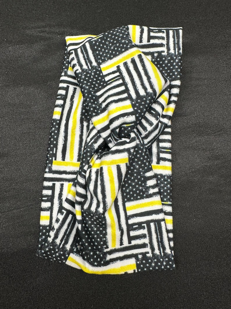 May include: A black and white headband with a yellow and black thin stripe pattern. The pattern is a repeated design of American flags with thin yellow stripes.
