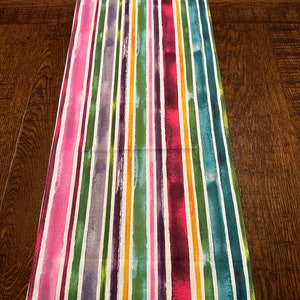 May include: A colorful table runner with vertical stripes in shades of pink, purple, green, yellow, and orange. The stripes are painted on a white background.