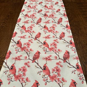 May include: A rectangular table runner with a repeating pattern of red cardinals perched on flowering branches. The background is a light cream color. The cardinals are a vibrant red with black accents, and the flowers are a soft pink.