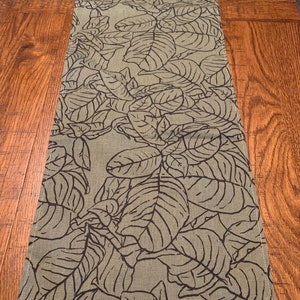 May include: A green table runner with a black leaf pattern. The runner is made of a linen-like fabric and has a subtle texture.