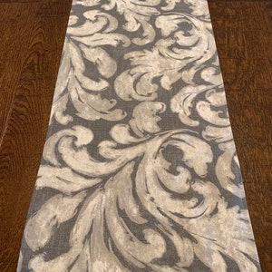 May include: A gray and white table runner with a damask pattern. The runner has a subtle sheen and a soft texture.