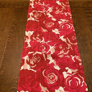 May include: A red and white table runner with a floral pattern of roses. The runner is made of a textured fabric.