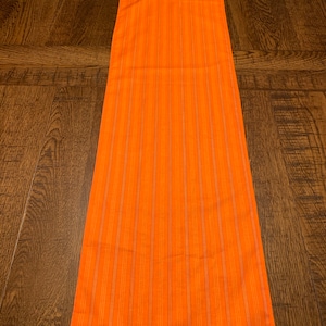 May include: Orange table runner with thin white stripes. The runner is made of a woven fabric and has a subtle texture.
