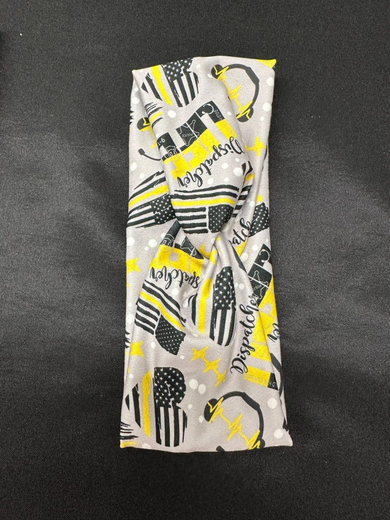 May include: Gray headband with a yellow and black pattern. The pattern includes the words "Dispatcher" and a thin yellow line flag. The headband is twisted.