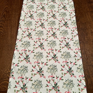 May include: A white table runner with a repeating pattern of green sprigs of mistletoe tied with red ribbons and red berries.