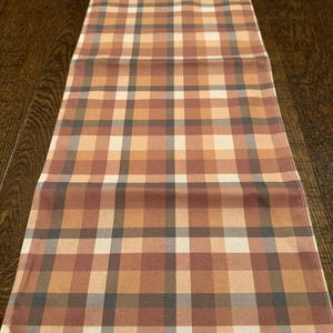 May include: A rectangular table runner with a plaid pattern in shades of brown, tan, and gray. The runner is made of fabric and is designed to be placed on a table. The plaid design is a classic pattern.