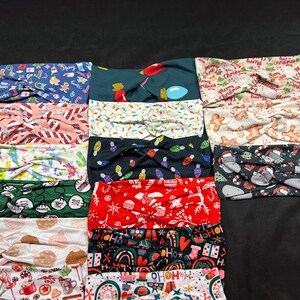 May include: A stack of 14 Christmas-themed headbands with various patterns and colors, including red, green, white, black, and brown. The headbands feature designs such as snowflakes, reindeer, candy canes, and Christmas lights.