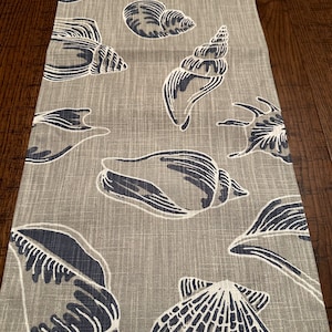 May include: A beige table runner with a repeating pattern of blue and white seashells. The seashells are in various shapes and sizes, including clams, conch shells, and scallop shells.